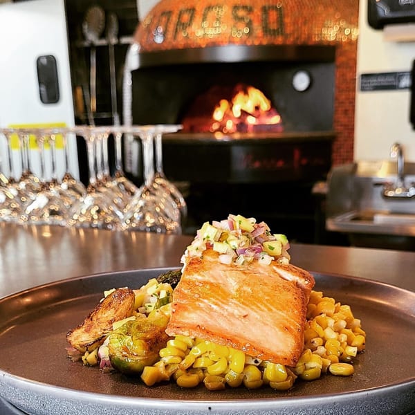 MOROSO's salmon dish with corn and brussels sprouts, with a wood-fired oven in the background.