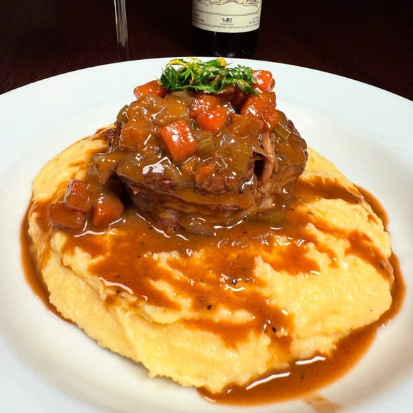 Osso buco with carrots and gravy over polenta, garnished with greens, on a white plate.