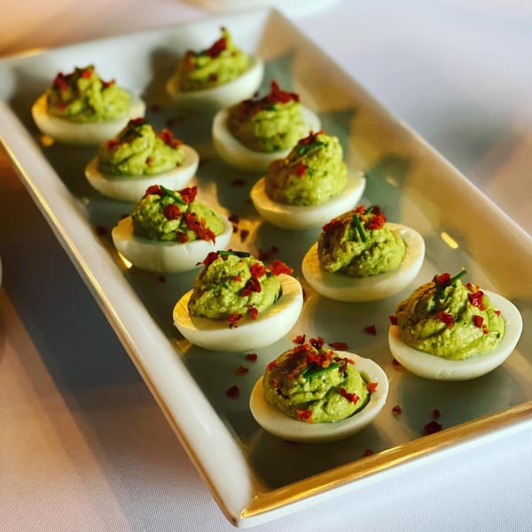 A white rectangular platter holds twelve deviled eggs with green filling and red pepper flakes.