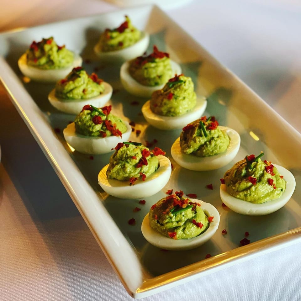 A white rectangular platter holds twelve deviled eggs with green filling and red pepper flakes.
