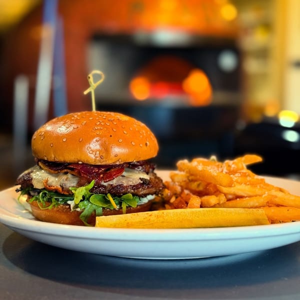 Gourmet burger with bacon, cheese, and arugula, served with fries, in front of a pizza oven.
