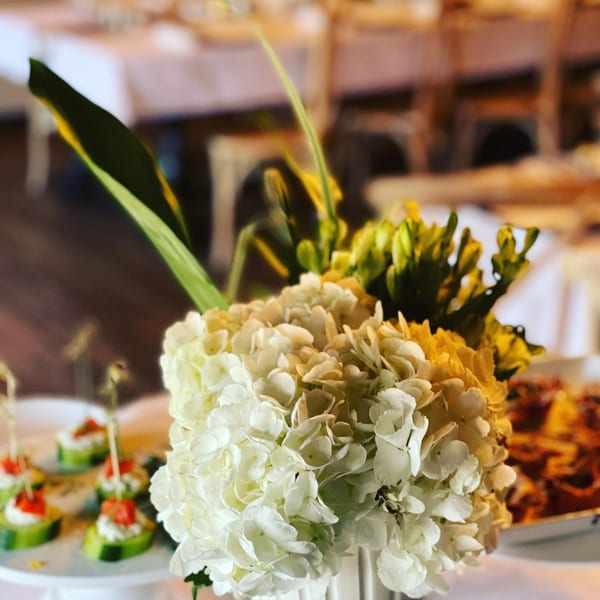 White and yellow floral centerpiece with cucumber appetizers and Italian food in the background.