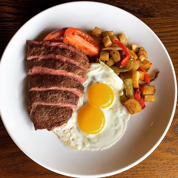 Sliced steak, two fried eggs, roasted potatoes, and tomato wedges on a white plate.