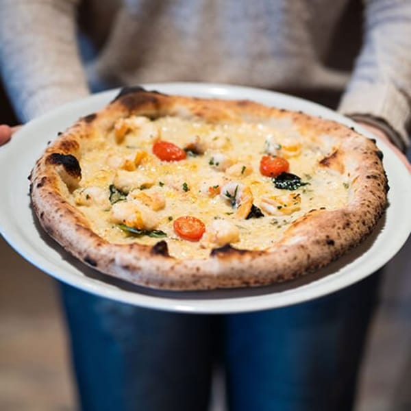 Hands holding a white plate with a seafood pizza topped with shrimp, cherry tomatoes, and black olives.