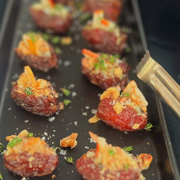 Close-up of stuffed dates with nuts, cheese, and herbs on a black tray with tongs.