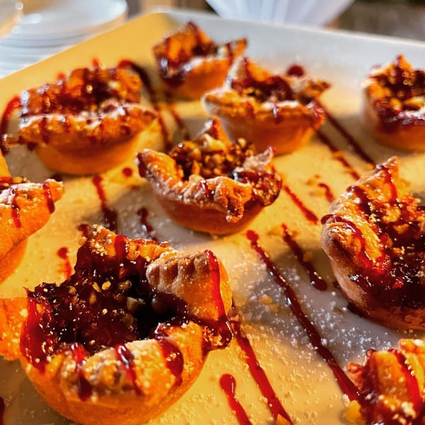 Mini Italian pastries with dark filling, red drizzle, and powdered sugar on a white plate.