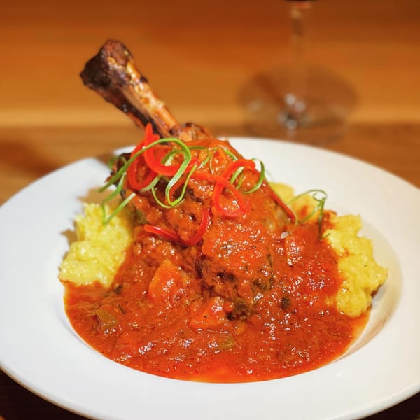 Braised lamb shank with red sauce, garnished with peppers and green onions, served with polenta.