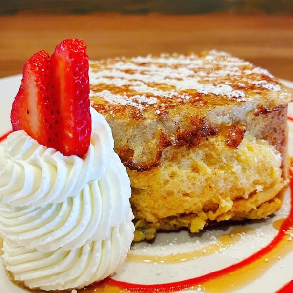Thick-cut French toast with powdered sugar, syrup, whipped cream, and two strawberry slices on a white plate.