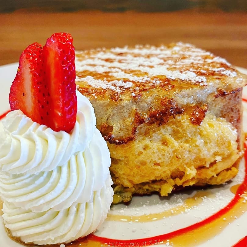 Thick-cut French toast with powdered sugar, syrup, whipped cream, and two strawberry slices on a white plate.